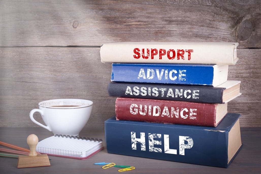 A stack of five books labeled with "SUPPORT," "ADVICE," "ASSISTANCE," "GUIDANCE," "HELP," next to a cup of coffee, a small notepad, colorful paper clips, and pencils on a wooden surface.